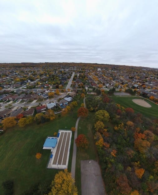 An aerial view of a suburban neighborhood in the United States with well-kept lawns and sidewalks, with several houses displaying the American flag, set against a backdrop of a sunny sky.