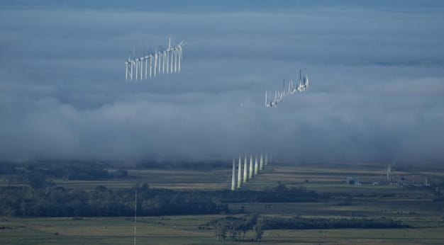 Wide shot of a military installation featuring several large wind turbines against a backdrop of rolling hills. The turbines are actively spinning, and the scene conveys a sense of clean energy production in a secure military environment.